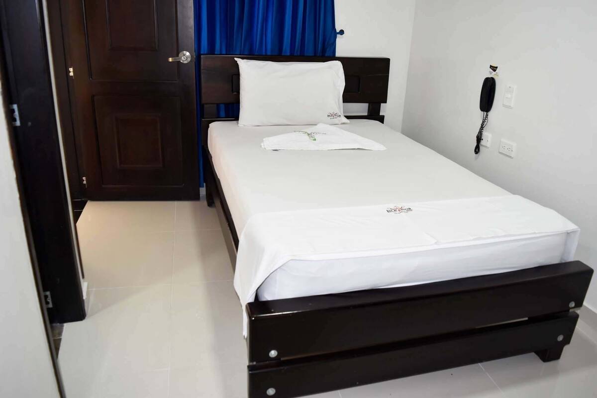 Una cama individual con colchón blanco y cabecera negra en una habitación con puerta cerrada y cortina azul.