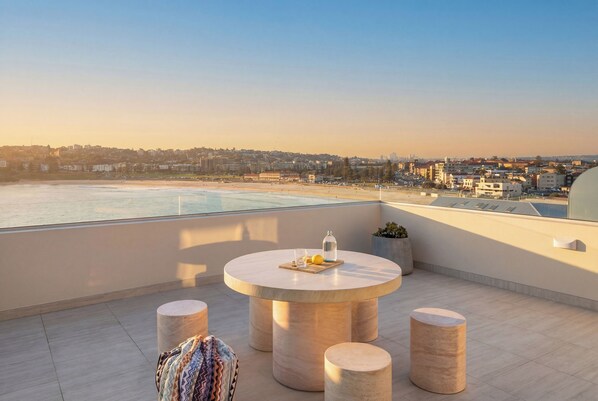 A rooftop terrace with a round table, four stools, and a view of the beach and cityscape.