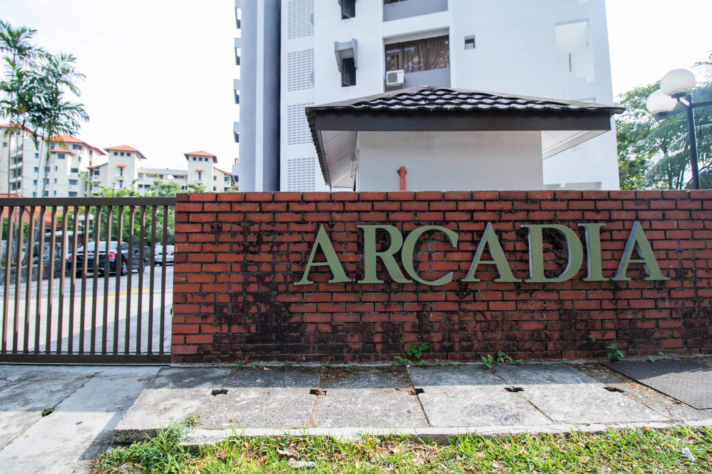 A brick wall with the name "ARCADIA" in large letters, a gated entrance, and a modern building in the background.