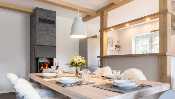 Un espace de repas moderne avec une longue table en bois, dressée pour quatre personnes, une cheminée en pierre et un luminaire suspendu.