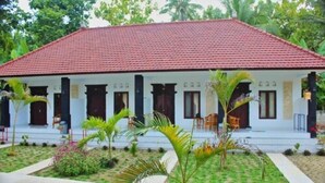 A two-story building with a red-tiled roof and white walls. There are multiple windows and doors, and the building is surrounded by a garden with palm trees and other greenery.