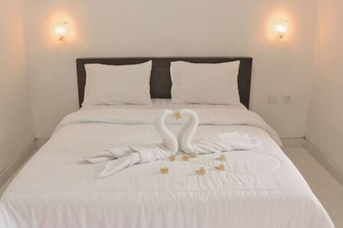 A neatly made bed with a white duvet and pillows, two wall-mounted lights, and a heart-shaped towel arrangement on the bed.