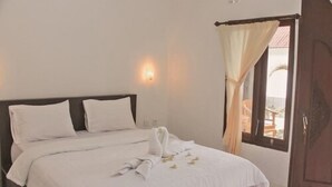 A neatly made bed with white linens and a towel arrangement, a wall-mounted light fixture, and a view through a window with curtains.