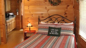 Een knus slaapkamer in een chalet met een houten plafond, een bed met beddengoed met een patroon, een nachtkastje met een lamp, en een keuken die zichtbaar is door een open deur.