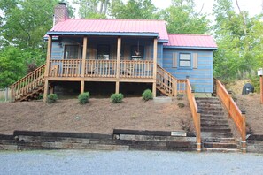 Une maison en bois à deux étages, avec un toit rouge, une terrasse en bois et un escalier menant à l’entrée.