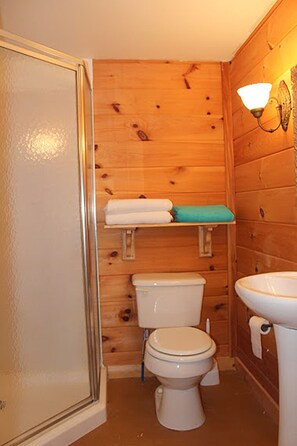Une petite salle de bain aux murs lambrissés, équipée de toilettes, d’un lavabo et d’une étagère où sont rangées les serviettes.