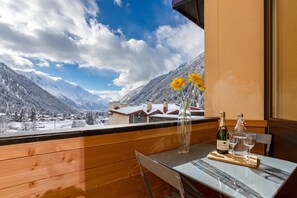 A table with a bottle of champagne, glasses, and a vase with flowers on a balcony overlooking a snowy mountain landscape.