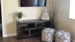 Ein Wohnzimmer mit einem dunklen Holz-Entertainment-Center, einem darüber montierten Flachbildfernseher, einer kleinen Topfpflanze, einer Buddha-Statue und zwei strukturierten Hocker.