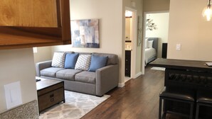 Ein modernes Wohnzimmer mit grauem Sofa, einem Couchtisch und einem Teppich. Ein Holzesstisch mit Barhocker steht im Raum. Der Boden ist mit Parkett ausgelegt und es gibt eine Deckenleuchte.