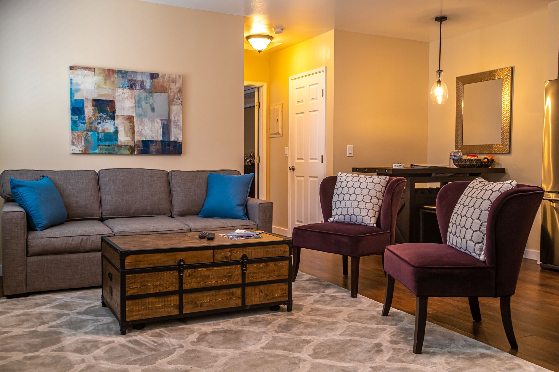 A living room with a gray sofa, a wooden coffee table, and a purple armchair. There is a painting on the wall and a mirror above the armchair.