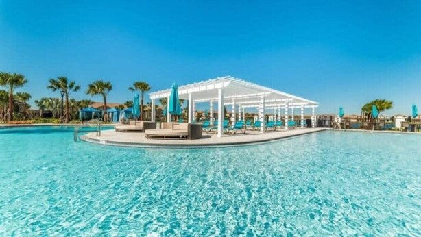 Une piscine d’un bleu éclatant, agrémentée d’une pergola blanche et de chaises longues, le tout entouré de palmiers.