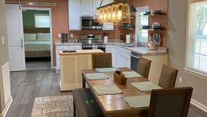 Een praktische keuken met een centraal eiland, een eetgedeelte met een houten tafel en stoelen, en een bed dat in de aangrenzende kamer zichtbaar is.