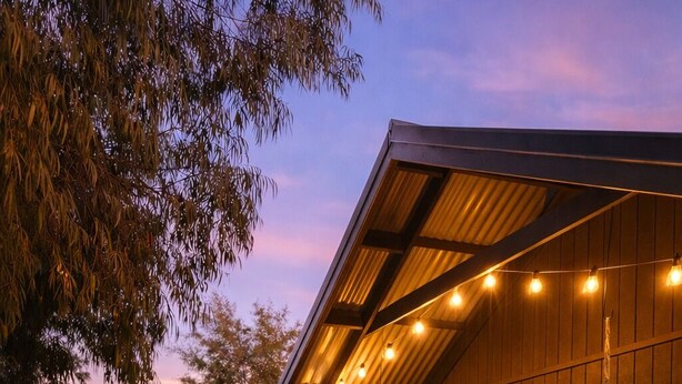 A wooden cabin with a covered porch, string lights, and a bed with pillows.