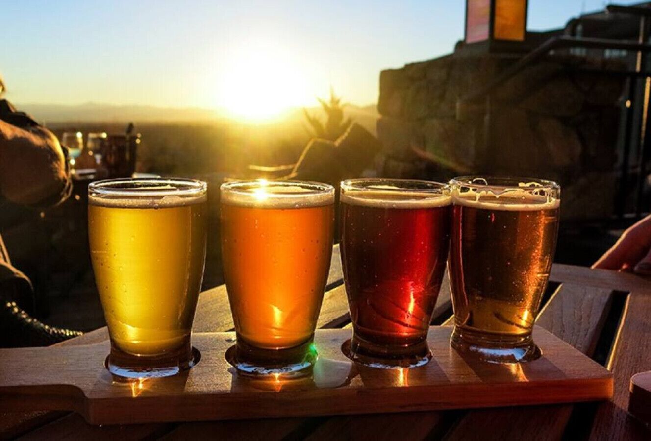 Four glasses of beer on a wooden tray with a sunset in the background.