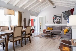 Uma sala de estar ampla com uma mesa de jantar, cadeiras, sofá e mesa de centro. Há uma janela grande com cortinas, uma lareira e uma televisão fixada na parede.