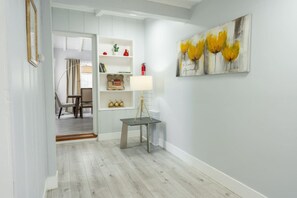 Un couloir avec un plancher en bois, un mur blanc, un tableau représentant des fleurs jaunes et une petite table avec une lampe.
