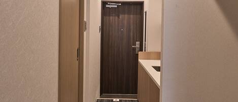 Un pasillo estrecho con una puerta de madera al final, una pequeña zona de lavamanos y un suelo con textura.