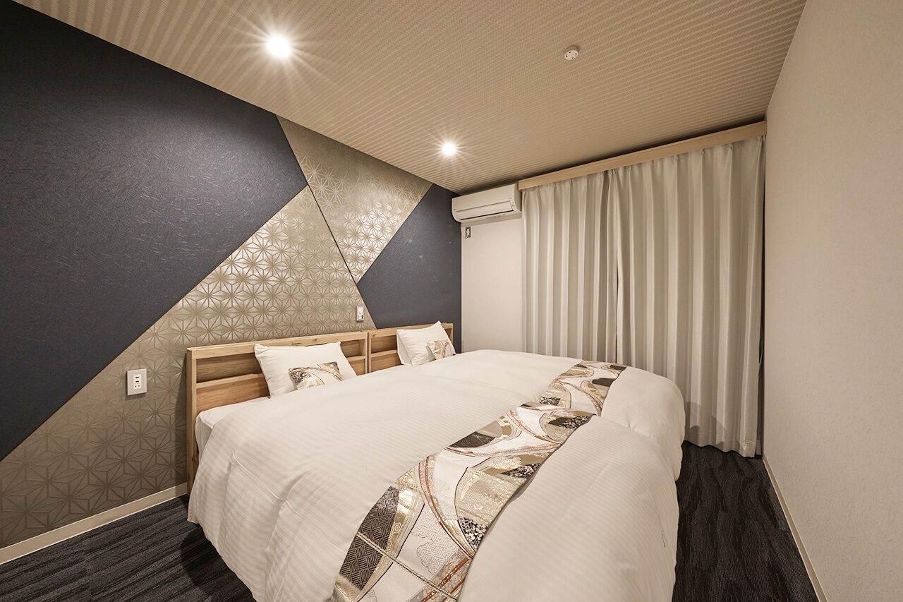 Una habitación de hotel con una cama, cabecera, pared con patrones geométricos, ventana con cortinas e iluminación empotrada en el techo.