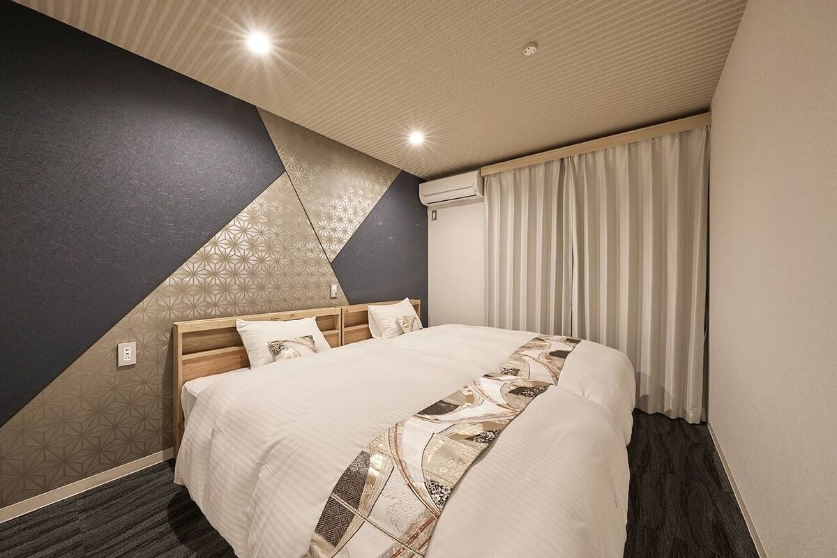 Une chambre d’hôtel avec un lit, une tête de lit, un mur orné de motifs géométriques, une fenêtre avec des rideaux et un plafond doté d’un éclairage encastré.