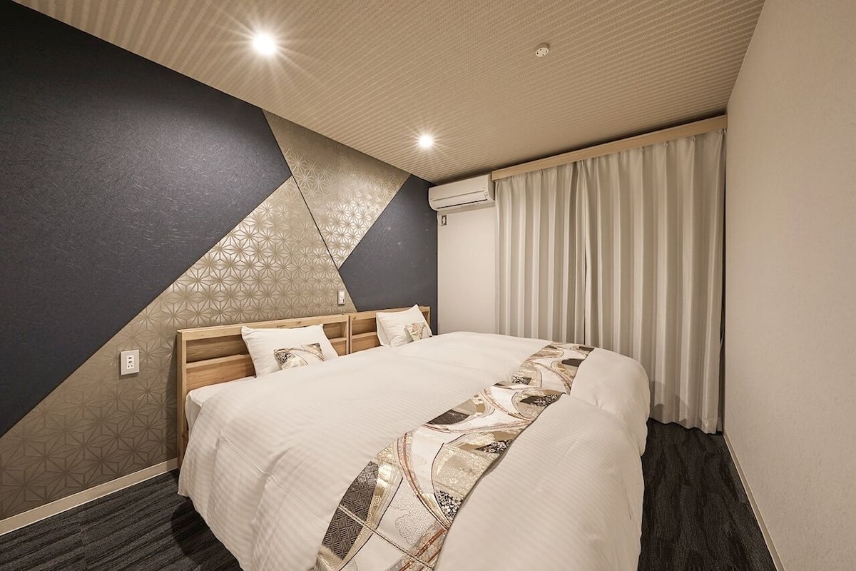Una habitación de hotel con una cama, cabecera, pared con patrones geométricos, ventana con cortinas e iluminación empotrada en el techo.