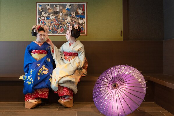 Deux personnes, vêtues de tenues traditionnelles, sont assises sur un banc, et un parapluie violet est posé à côté d’elles.