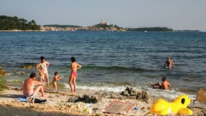 A beach with people swimming and sunbathing, a lighthouse in the background, and a rocky shoreline. - Holiday apartment with garden and terrace (Rovinj)