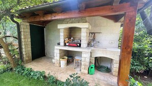 A stone-built outdoor kitchen with a wooden pergola overhead, a brick wall, and a green hose on the ground. - Holiday apartment with garden and terrace (Rovinj)