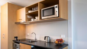 A compact kitchen with wooden cabinets, a stainless steel microwave, a kettle, and a fruit bowl on the counter. - Quest Tauranga Central (Tauranga)