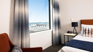 A hotel room with a large window, a bed, a bedside table with a lamp, and a chair. - Quest Tauranga Central (Tauranga)