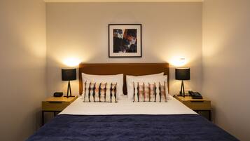 Ein Hotelzimmer mit einem großen Bett, zwei Nachttischen mit Lampen, einem gerahmten Kunstwerk über dem Bett und einer dunkelblauen Tagesdecke.