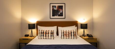 Ein Hotelzimmer mit einem großen Bett, zwei Nachttischen mit Lampen, einem gerahmten Kunstwerk über dem Bett und einer dunkelblauen Tagesdecke.