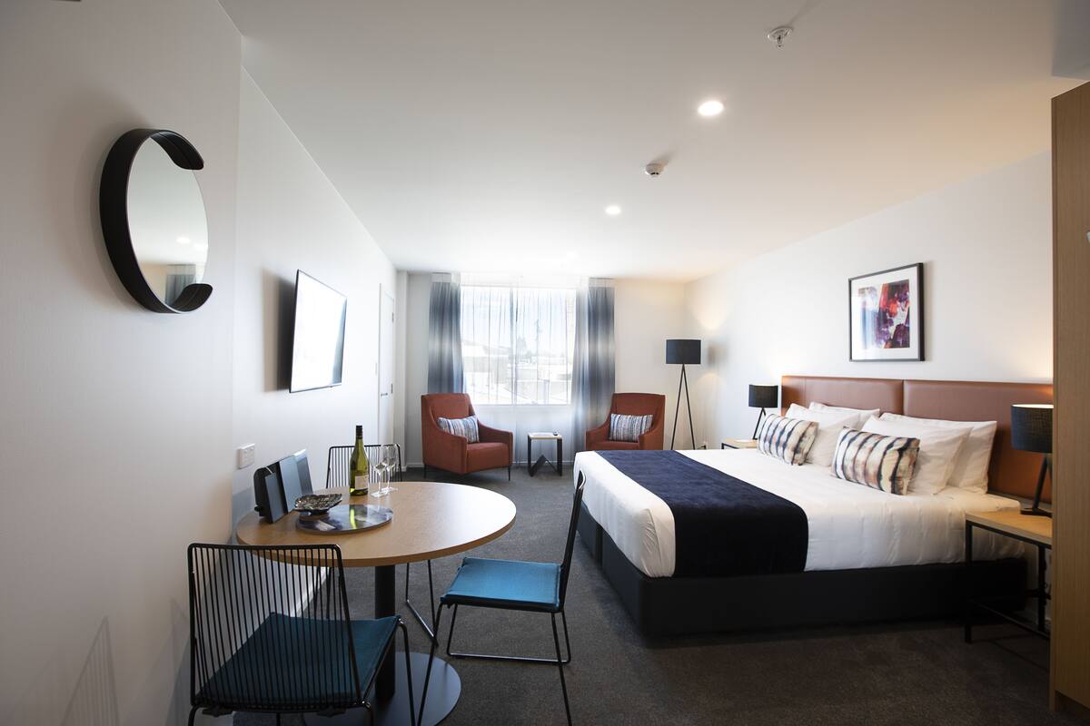 A hotel room with a bed, two armchairs, a table with a bottle and glasses, a TV, and a large mirror.