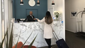 A hotel reception desk with a clock, a vase of flowers, and a sign that reads "QUEST". - Quest Tauranga Central (Tauranga)