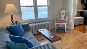 Olohuone, jossa on sohva, sohvapöytä, tuoli, lamppu, patteri ja ikkunasta avautuva merinäköala.