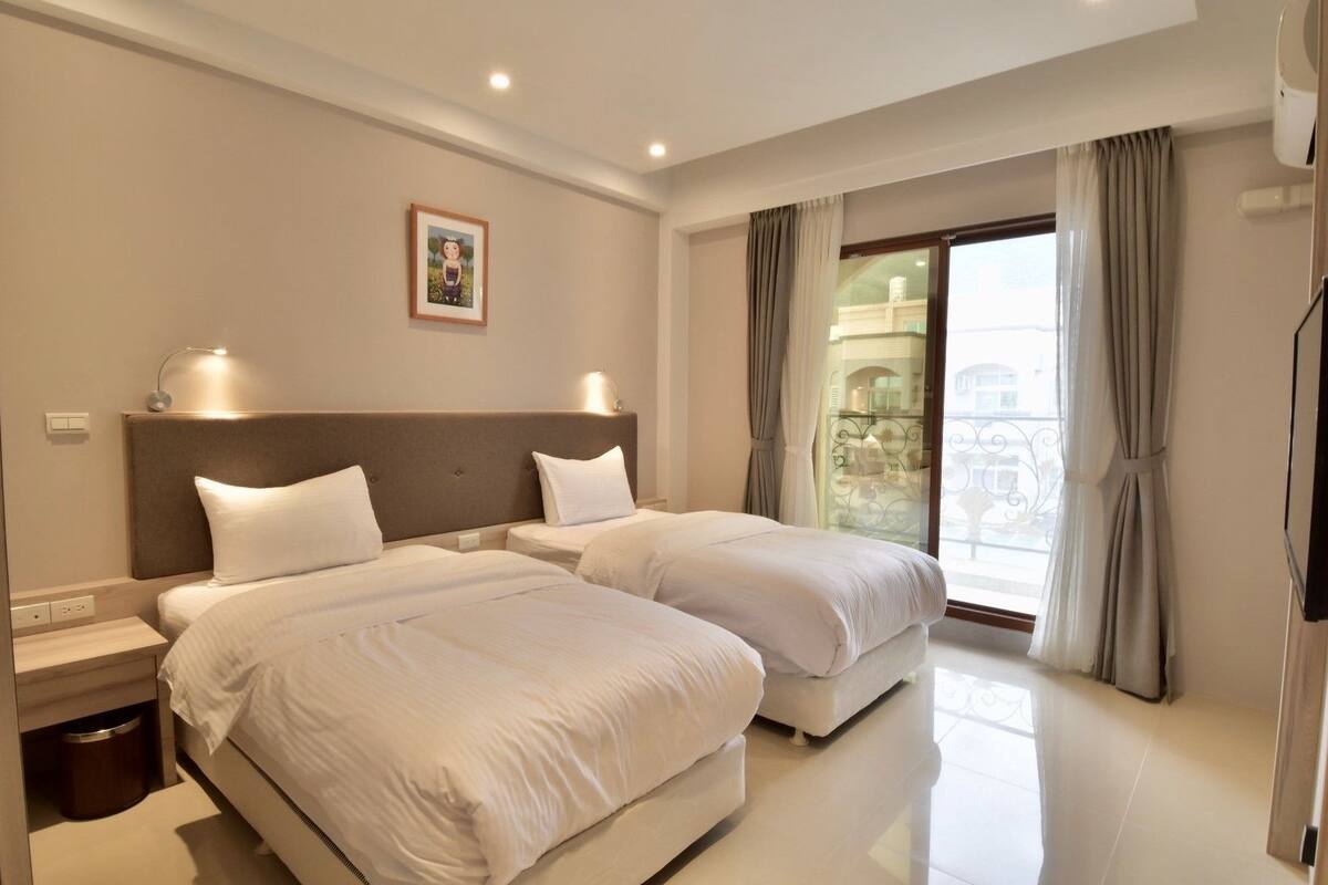 Habitación de hotel con dos camas, un ventanal con cortinas, un cuadro enmarcado en la pared y un cubo de basura debajo de la cama.