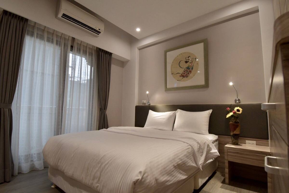 Una habitación de hotel con una cama grande, una mesita de noche con un jarrón de flores, un equipo de aire acondicionado y una obra de arte enmarcada en la pared.