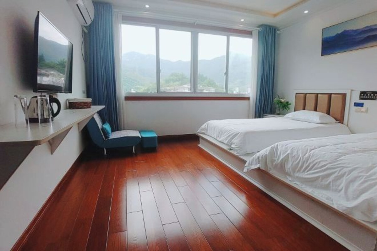 Habitación de hotel con piso de madera, dos camas, un televisor, un escritorio y ventana con vista a las montañas.