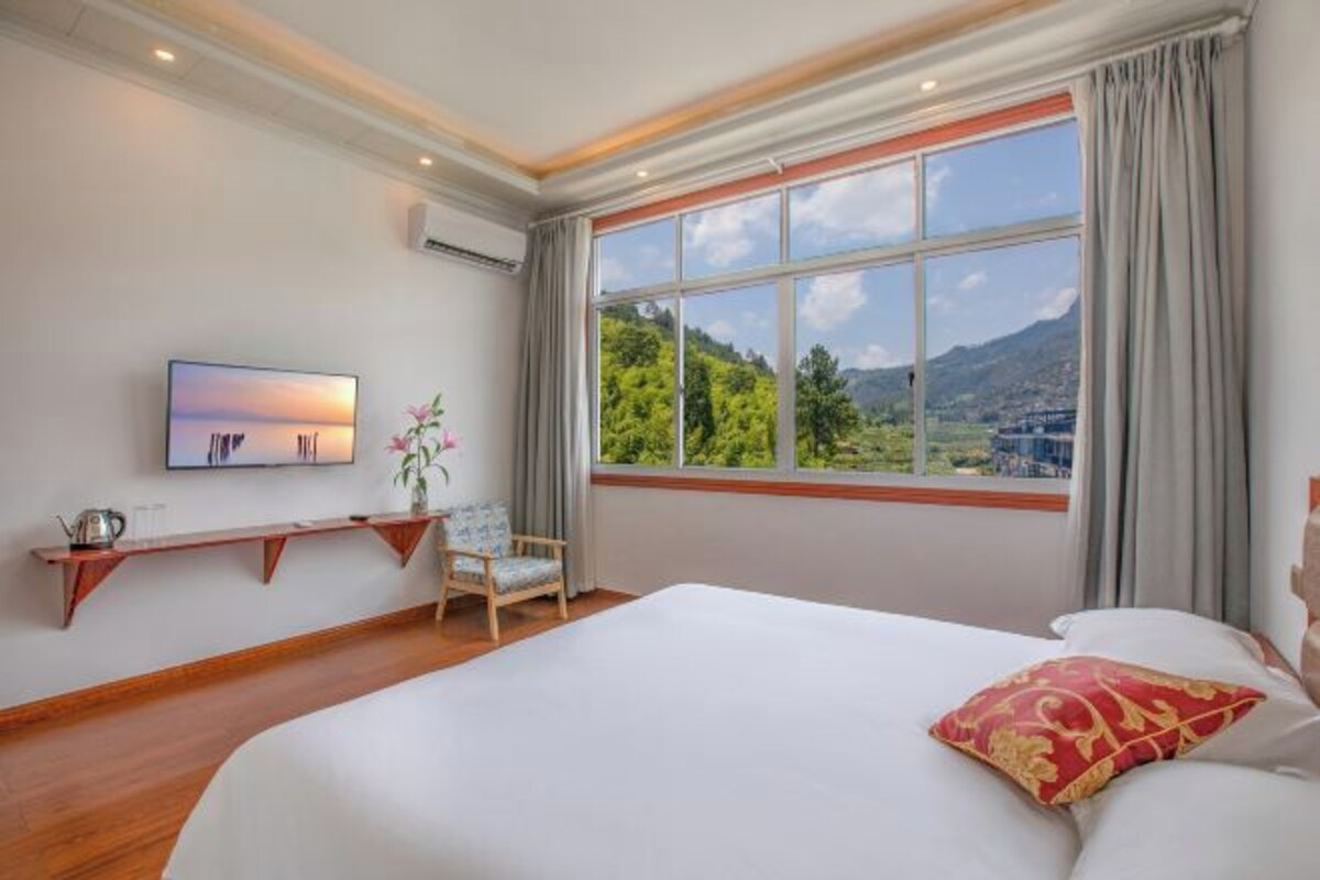 Habitación de hotel con una cama grande, un televisor montado en la pared, una ventana con vistas a la vegetación y montañas, y un estante de madera con una planta.