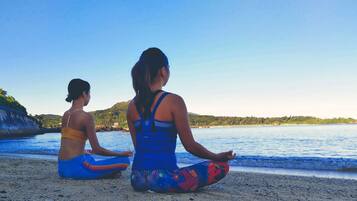 Duas pessoas meditando numa praia, com vista para o mar e colinas ao fundo.