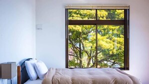 A bedroom with a large window, a bed, a bedside table, and a lamp. - The Little Garden (Miyazaki)