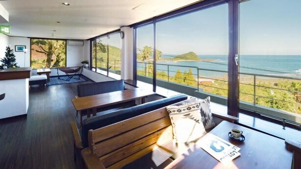A modern room with large windows, a wooden bench, a table with a cup, and a view of the sea and greenery. - The Little Garden (Miyazaki)