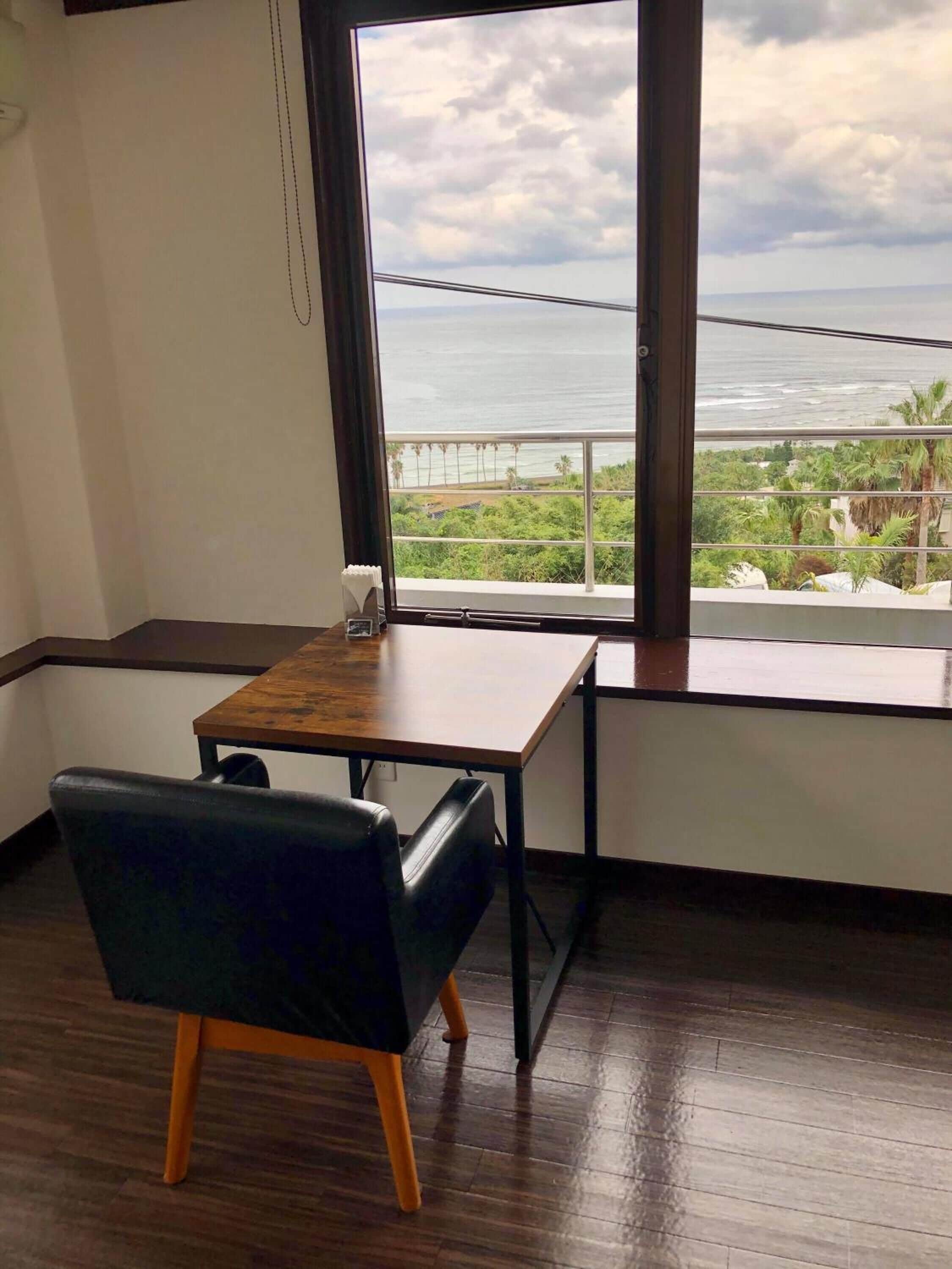 Una habitación con una mesa de madera y una silla negra, un ventanal con vista al océano y palmeras.