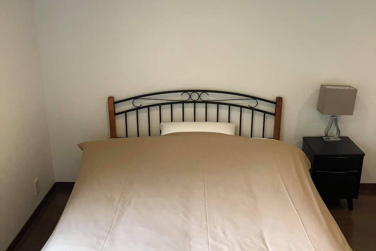 A bed with a beige mattress and a wooden headboard in a room with a white wall and a small nightstand with a lamp.