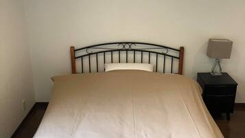 A bed with a beige mattress and a wooden headboard in a room with a white wall and a small nightstand with a lamp.