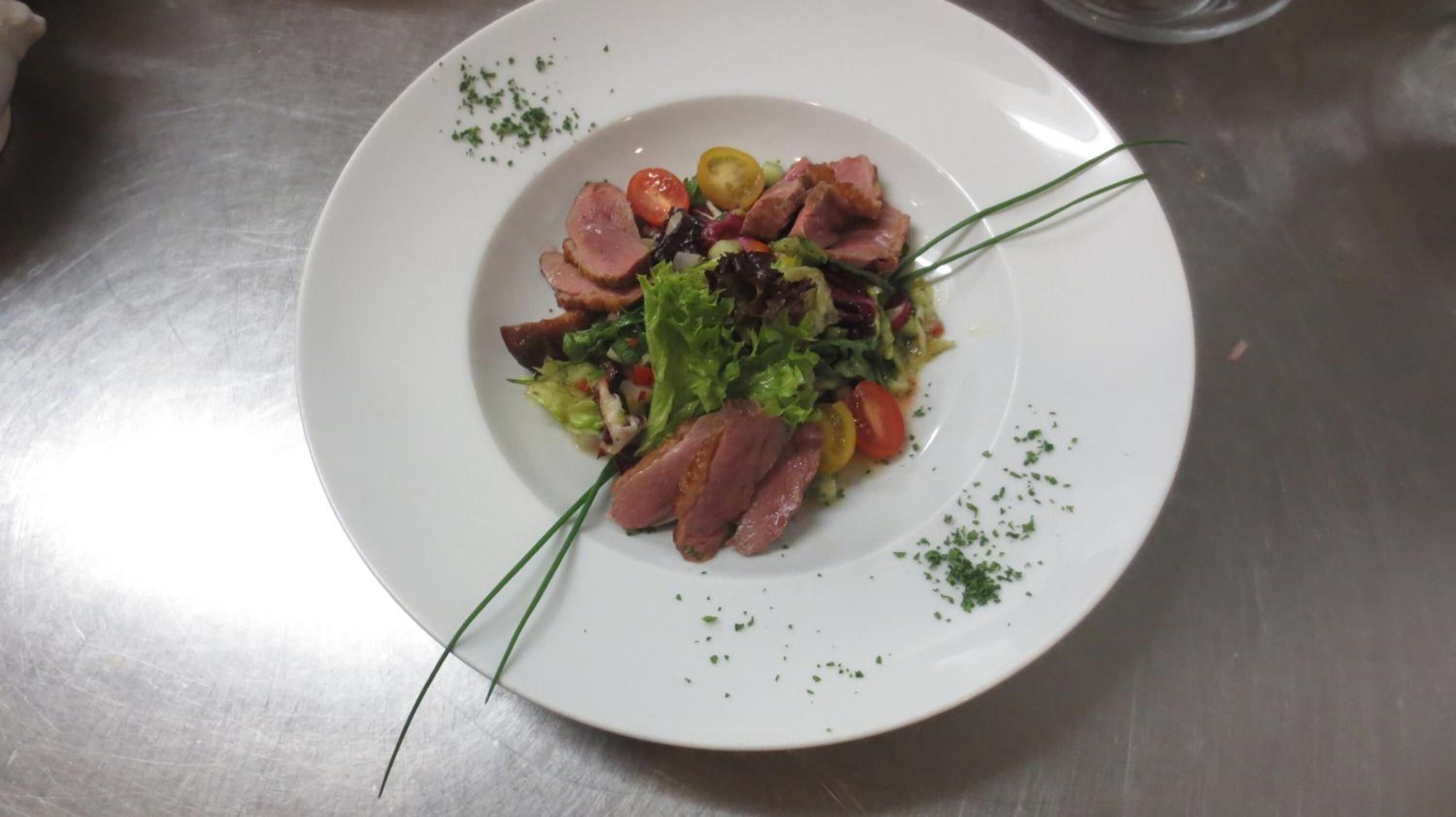 Un plato blanco con ensalada, carne en rodajas y guarniciones sobre una superficie metálica.