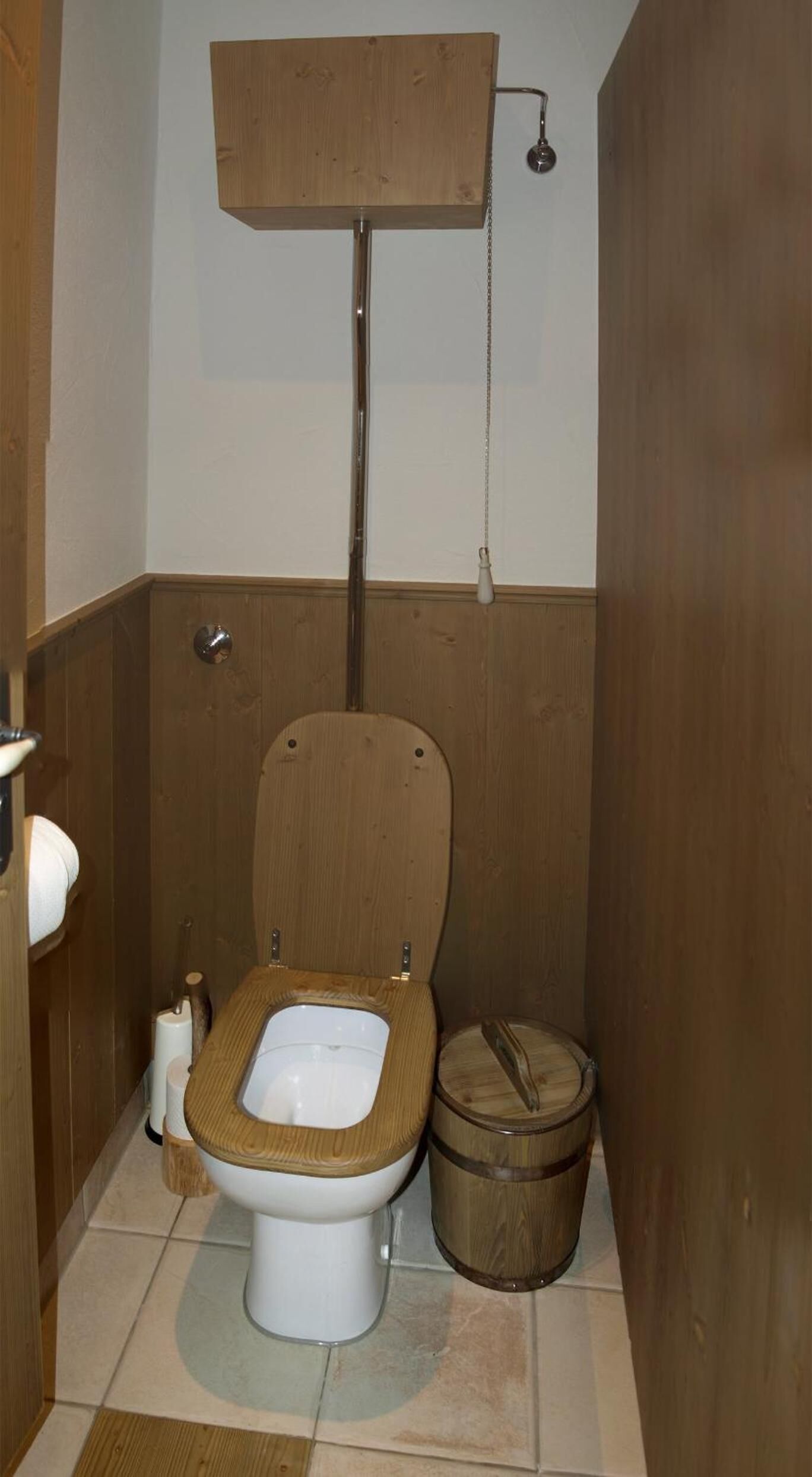 Baño pequeño con piso de madera, un inodoro, un balde de madera y un dispensador de papel higiénico.