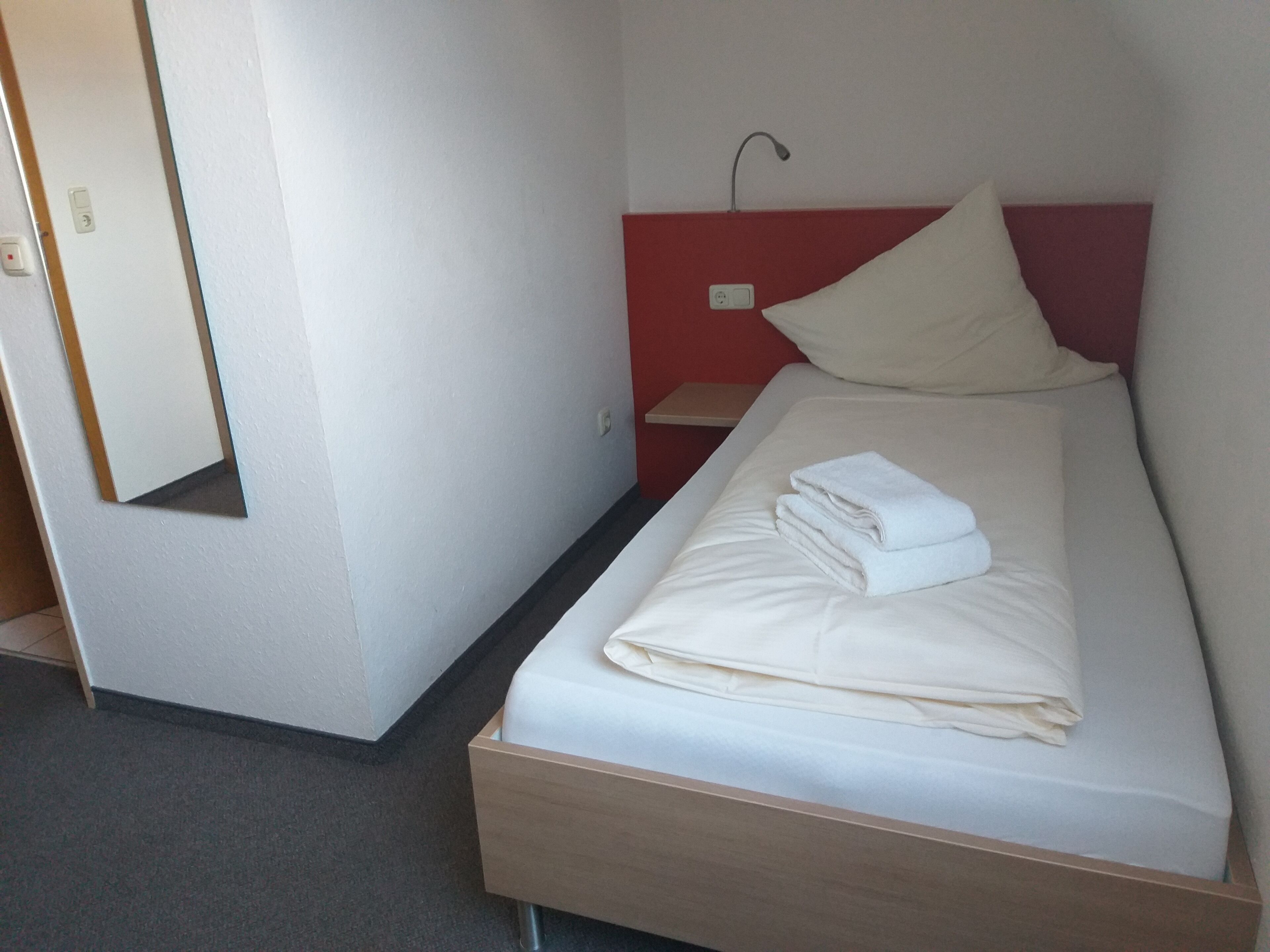 En pænt redt seng med et foldet hvidt håndklæde, et sengebord med en lampe og et spejl på væggen.