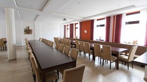 Une salle de conférence dotée d’une longue table et de nombreuses chaises, de grandes fenêtres ornées de rideaux rouges et d’un plafond blanc avec des luminaires encastrés.
