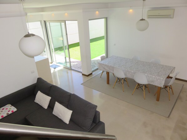 A modern living room with a grey sofa, a dining table with white chairs, and a large window with a view of a green lawn. - Alfarim House - close to Meco beach and Sesimbra (Alfarim)