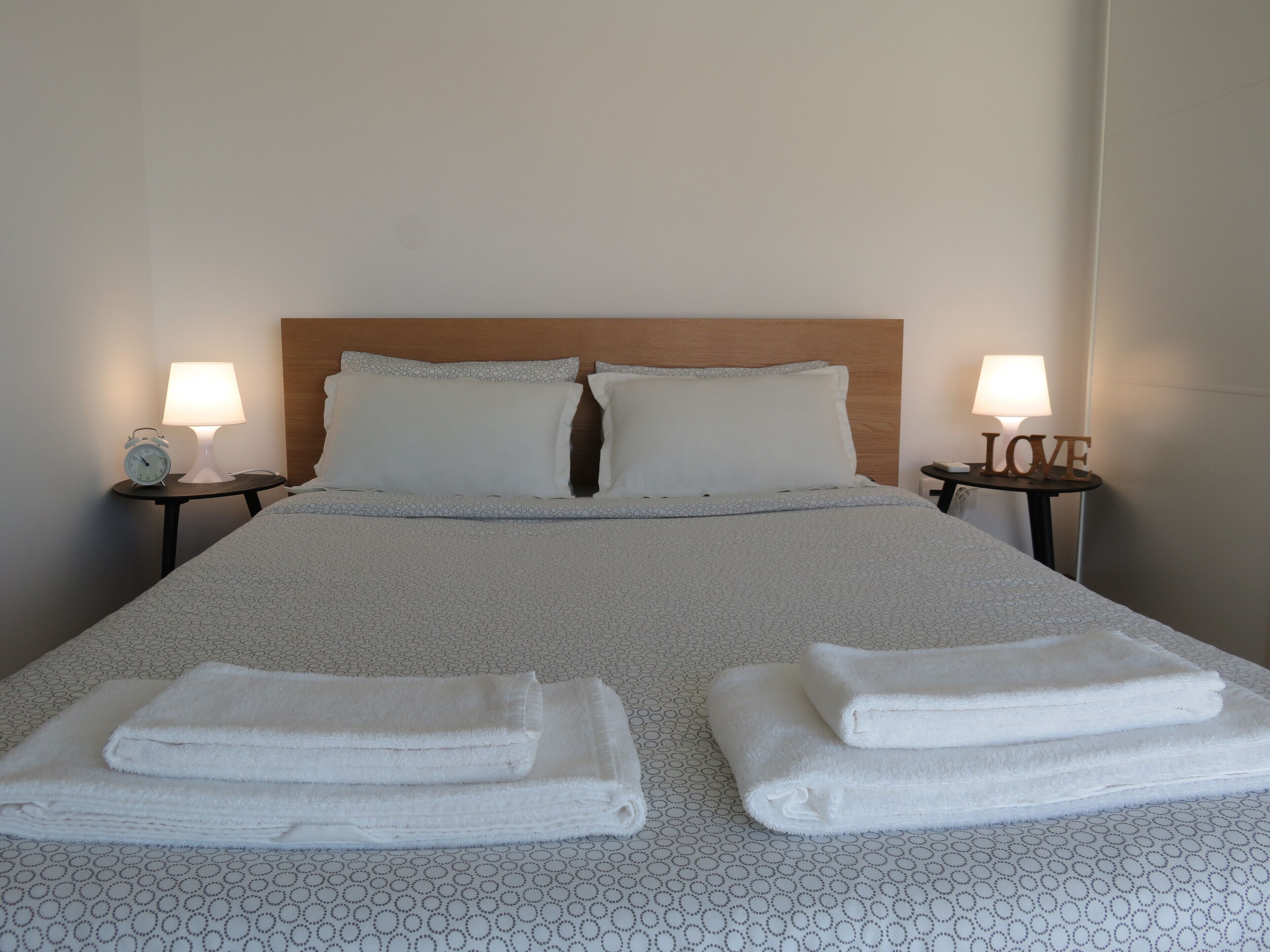Una cama bien hecha con una colcha estampada, dos almohadas y dos mesitas de noche con lámparas y un reloj en un lado.
